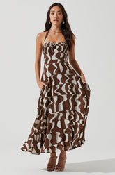 A woman is wearing a sleeveless, maxi dress with a square neckline and thin straps. The dress features a bold brown and white abstract pattern with swirling, wavy shapes throughout. She is also wearing nude, open-toe high heels.